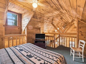 A wooden room with a sloped ceiling, a bed with a patterned blanket, a television on a stand, and a white chair.