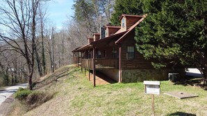 A rustic cabin with a porch and a stone foundation, surrounded by trees and a dirt path. - Country Charm A - Pet Friendly Group Cabin (Helen)