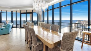 A spacious dining area with a wooden table and wicker chairs, large windows with a city view, and a balcony with a view of the ocean.