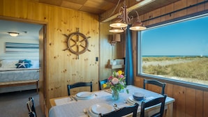 A wooden cabin with a dining area featuring a table set for four, a ship's wheel on the wall, and a view of the beach through a large window.