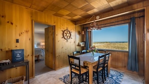 A wooden interior with a dining table, chairs, a ship's wheel, and a view of the beach.
