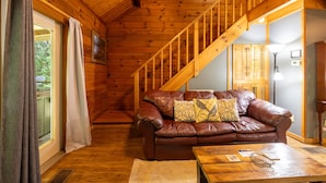 A wooden interior with a brown leather sofa, a wooden staircase, and a view through a glass door. - Love Bird - The Perfect Nest! (Sautee Nacoochee)