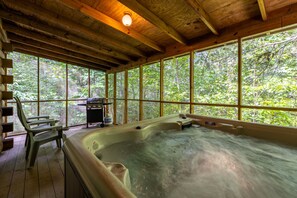 Un bain à remous dans un chalet en bois doté de grandes fenêtres et d’un barbecue à l’extérieur.