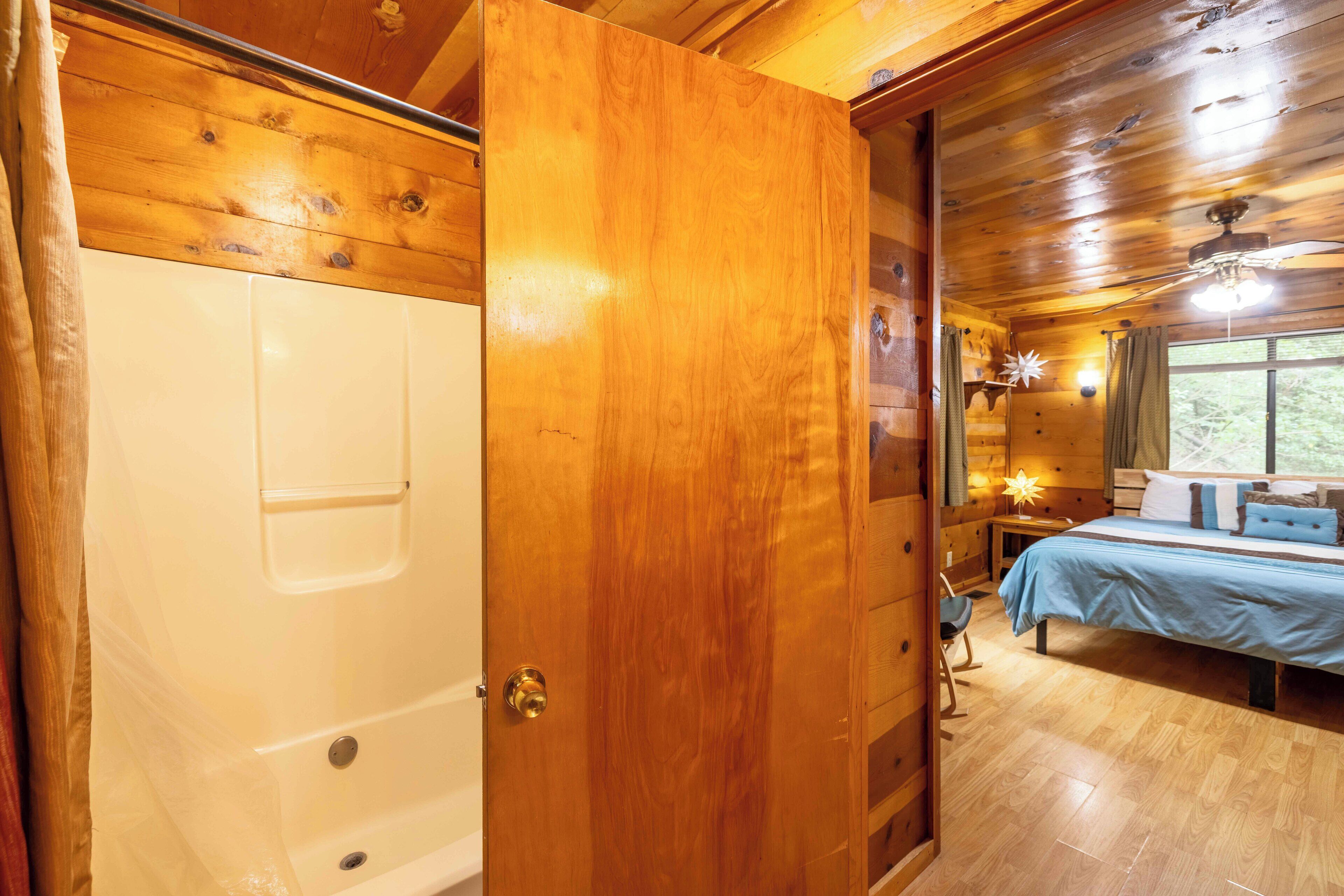 A wooden cabin interior with a wooden door leading to a bedroom with a bed, a nightstand, and a window.