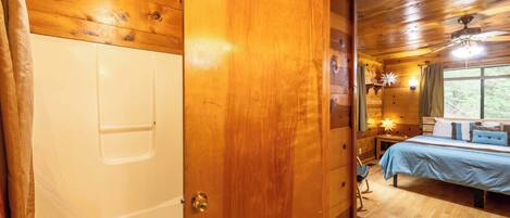 A wooden cabin interior with a wooden door leading to a bedroom with a bed, a nightstand, and a window.