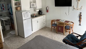 Eine kompakte Küche mit weißen Schränken, einer Mikrowelle und einer Kaffeemaschine. Es gibt einen kleinen Essbereich mit Korbstühlen und einem Tisch. Ein Fernseher ist an der Wand montiert, und ein Feuerlöscher ist sichtbar.
