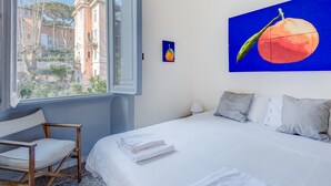 A hotel room with a bed, a chair, a window with a view of buildings, and a painting of a peach. - Charming, quiet, sunny two-bedroom in Trastevere with great view (Roma)
