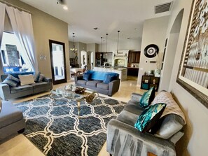 Un salón moderno con un sofá gris, una mesa de centro y un tapete estampado. Al fondo, se observa una zona de comedor con un sofá azul y una cocina.