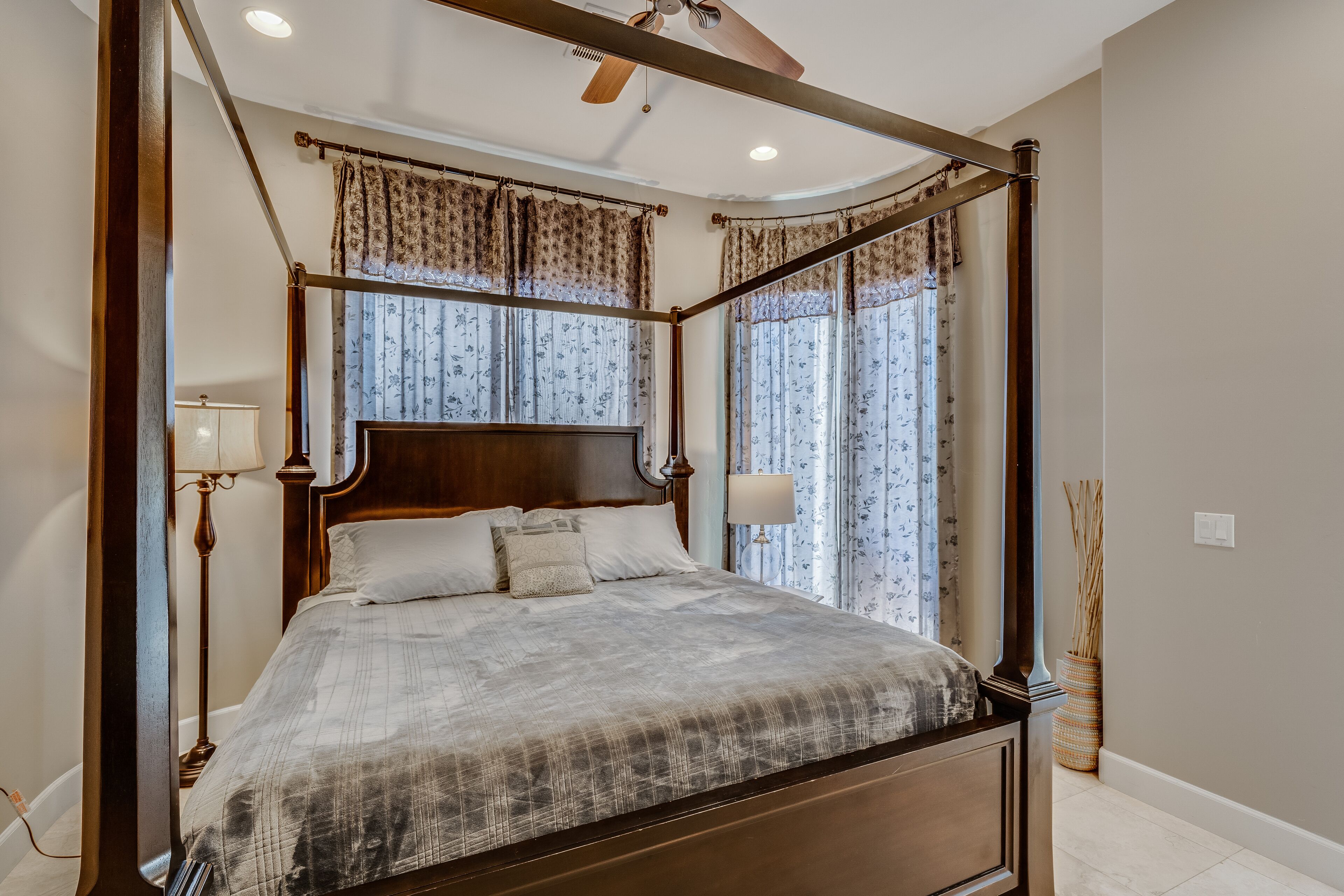 A four-poster canopy bed with a wooden headboard, a bedside table with a lamp, and a vase with reeds.
