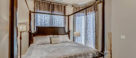A four-poster canopy bed with a wooden headboard, a bedside table with a lamp, and a vase with reeds.