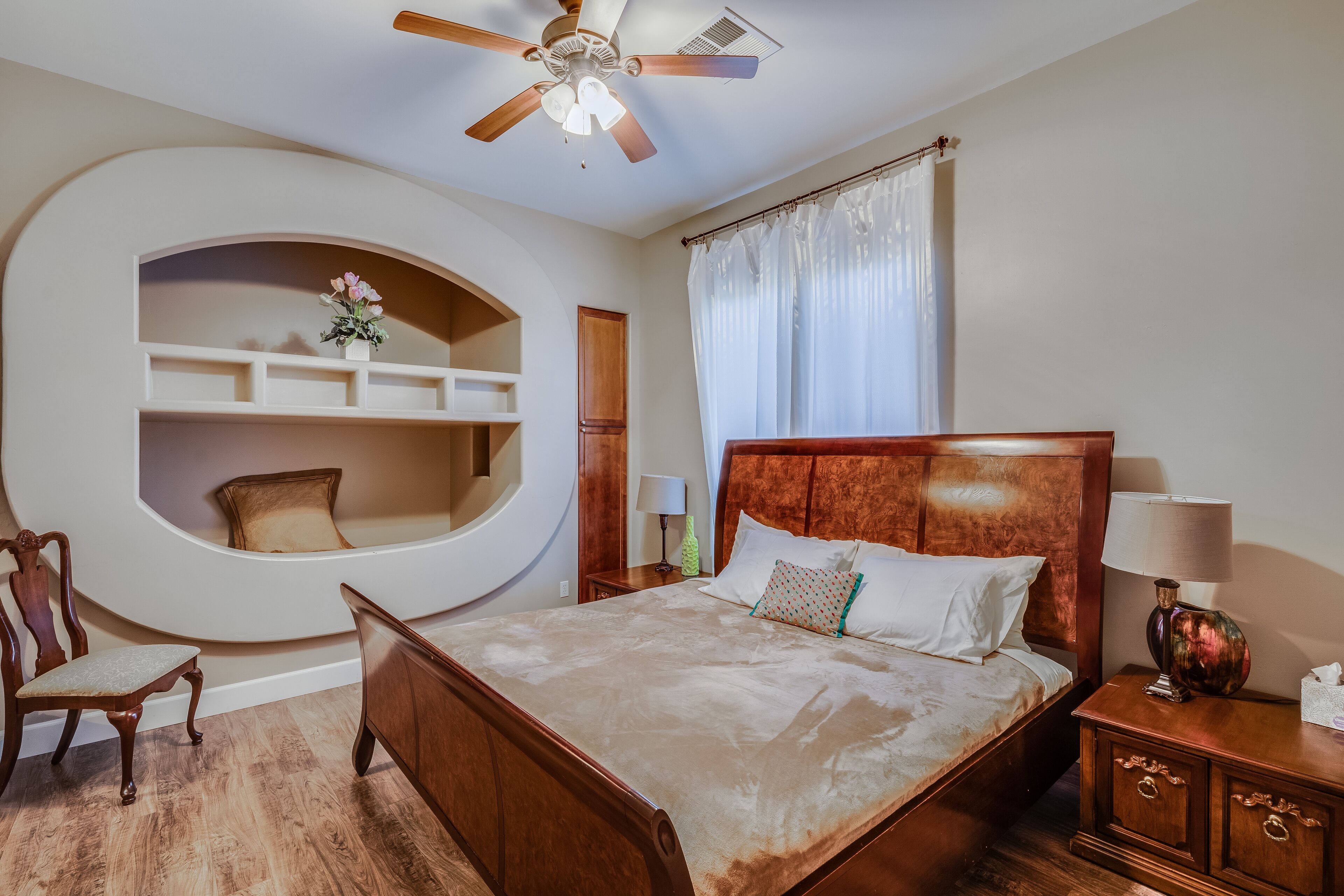 Une chambre avec un lit en bois, un ventilateur de plafond, une bibliothèque intégrée, une chaise et une table de chevet avec une lampe.
