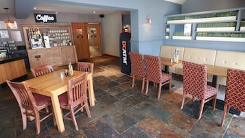 Una cafetería con mostrador de madera, zona de estar con sillas estampadas y suelo de baldosas de piedra.