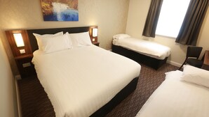 Een hotelkamer met een groot bed, een kleiner bed, een nachtkastje met lamp, een schilderij aan de muur en een raam met gordijnen.
