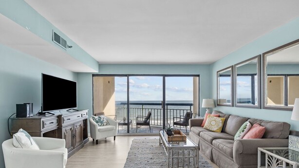 A spacious living room with a large TV, comfortable seating, a coffee table, and a view of the ocean through large windows.