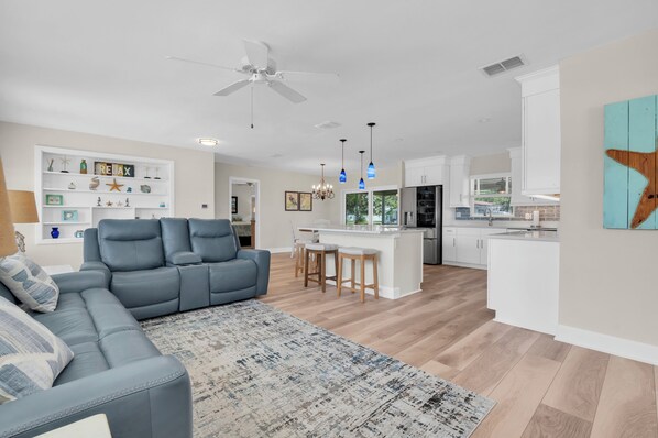 A modern living room with a blue sofa, a patterned rug, a ceiling fan, and a kitchen area with a bar. - Beautiful 3BD,2BA Waterfront Pool Home (Saint Petersburg)