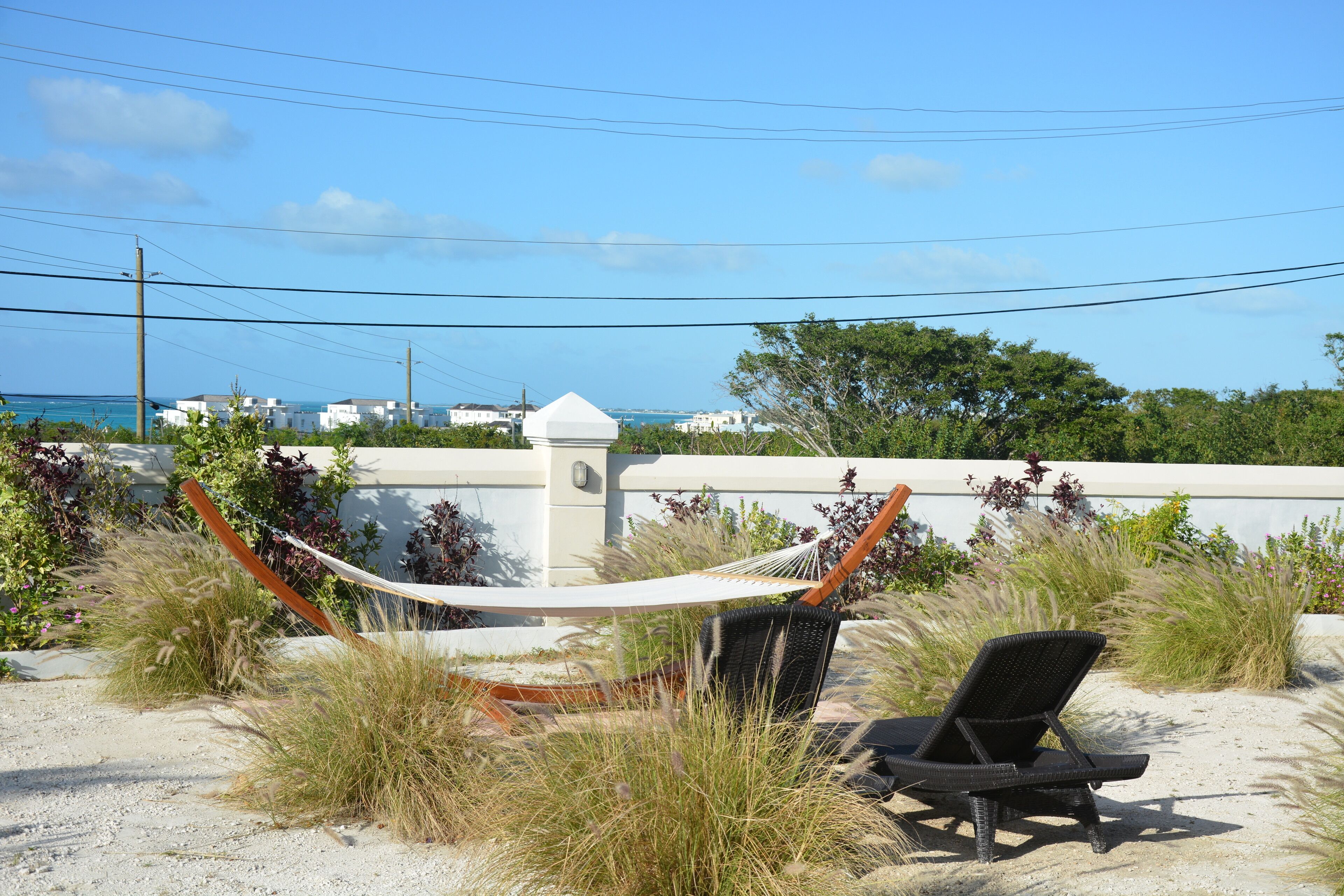 A hammock and a lounge chair on a sandy beach with a view of the ocean and buildings in the background.