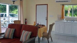 Una stanza con zona pranzo, divano, tavolo con bottiglia e bicchieri, estintore, finestra con vista sugli alberi e angolo cottura.