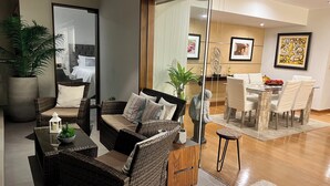 Un salón moderno con muebles de mimbre, un separador de vidrio y una zona de comedor con sillas y mesa blancos.