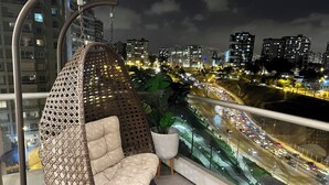 Un balcón con una silla colgante, una planta en maceta y vistas de la ciudad de noche.