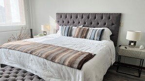 Una cama bien hecha con un cabecero gris con botonadura, ropa de cama blanca y mantas a rayas.