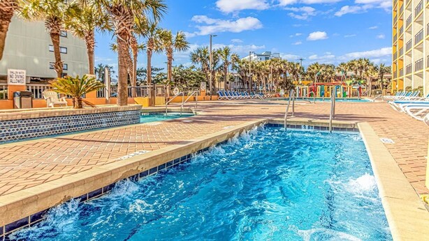 Une piscine bordée de palmiers, avec des chaises longues et un ciel d’un bleu limpide.