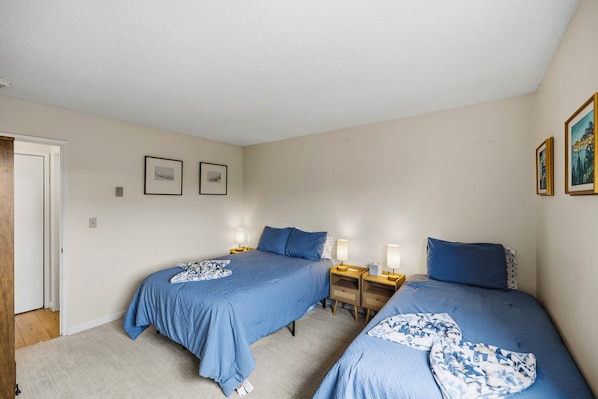 Un dormitorio con dos camas, cada una con colcha azul y manta estampada. Hay mesitas de noche con lámparas y una puerta a la izquierda.