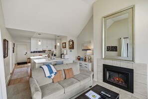 A modern living room with a fireplace, a sofa, a coffee table, and a kitchen in the background.