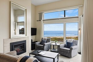 Un salón con chimenea, asientos, un televisor de pantalla plana y un gran ventanal con vistas a la playa.