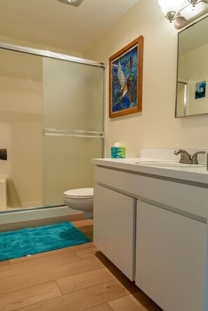 Ein Badezimmer mit einer Duschkabine aus Glas, einer weißen Toilette, einem Waschbecken mit Spiegel und einem gerahmten Kunstwerk an der Wand.