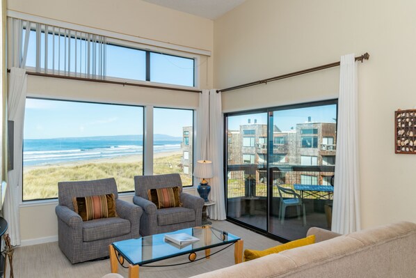Amplio salón con un gran ventanal con vistas a la playa, dos sillones, un sofá y una mesa de centro de cristal.