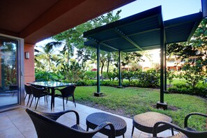 Un patio exterior con zona de estar cubierta, entrada por puerta de vidrio y abundante vegetación.
