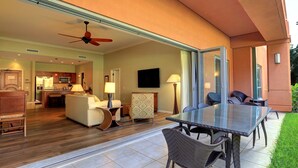 A modern hotel room with a glass door leading to a balcony, a dining area with a glass table, and a kitchen visible through the door.