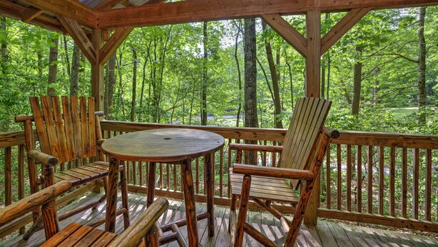 Une terrasse en bois avec une table ronde et deux chaises, entourée d’une balustrade en bois, donnant sur une zone boisée.