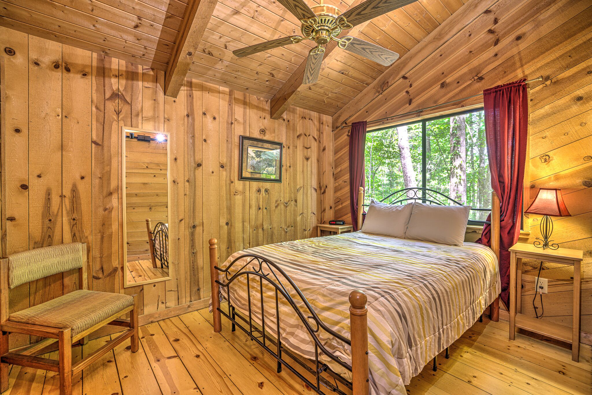A wooden cabin bedroom with a bed, a chair, a nightstand, a lamp, and a ceiling fan.