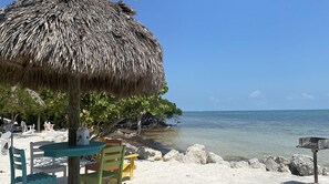 A beach scene with a thatched umbrella, a table with chairs, and a clear view of the ocean. - Paradise awaits, remodeled, marina, pickleball, tennis, pools, salt water lagoon (Key Largo)