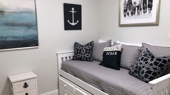 Ein Schlafzimmer mit einem Bett, einem Nachttisch, einer Schublade, einem gerahmten Bild von Surfern und einem Meeresblick-Gemälde.