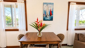 Un'area pranzo con un tavolo in legno, sedie in vimini, un vaso con fiori e un quadro raffigurante dei velieri.