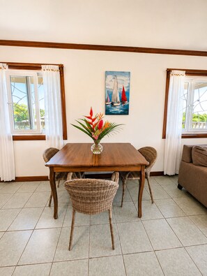 Een eetruimte met een houten tafel, rieten stoelen, een vaas met bloemen en een schilderij van zeilboten aan de muur.