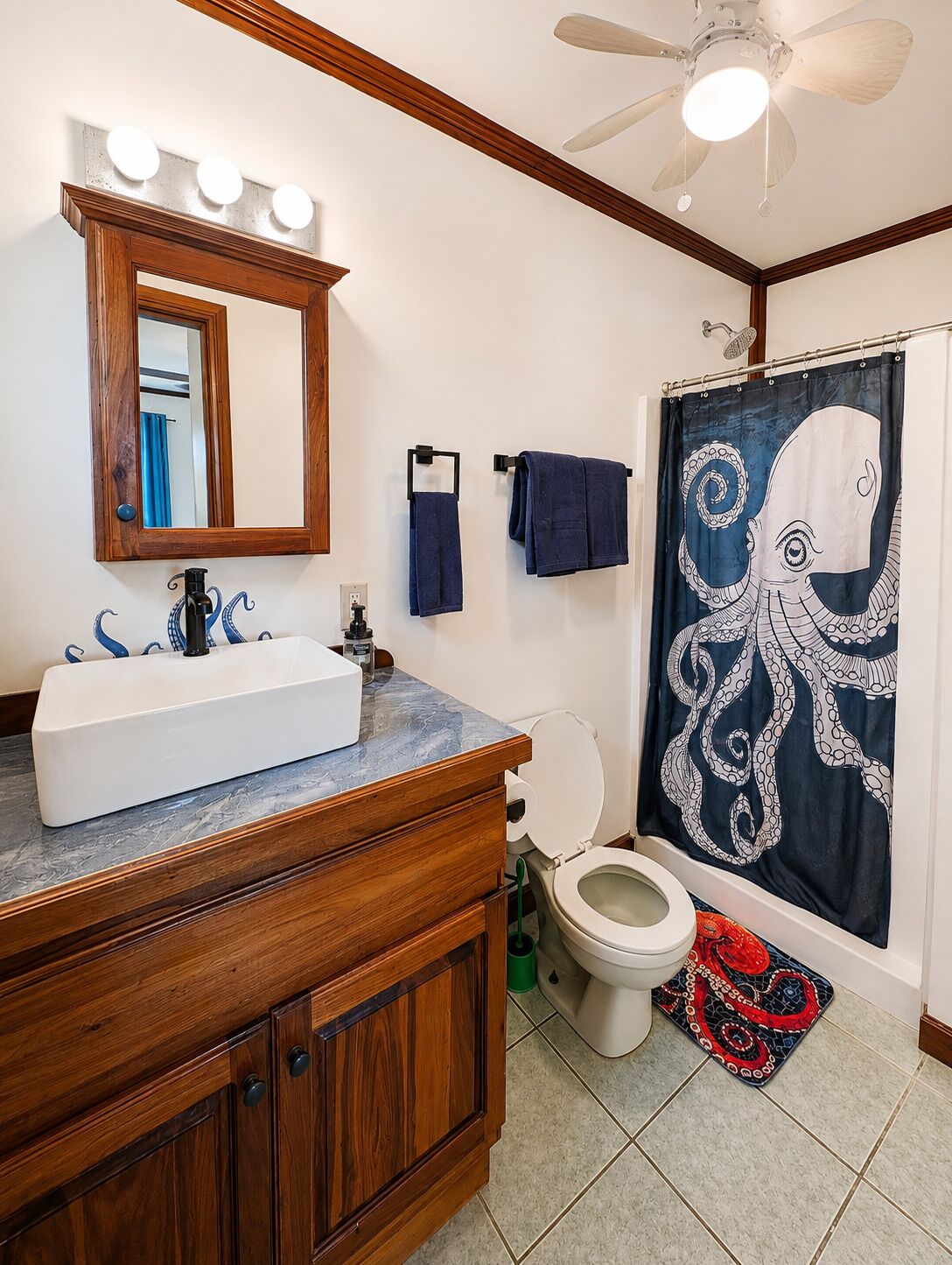Une salle de bain avec un meuble-lavabo en bois, un lavabo blanc, un miroir, un ventilateur de plafond et un rideau de douche à motifs de poulpes.