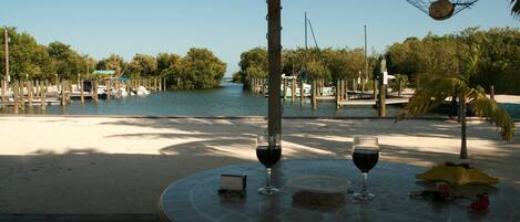 Une table ronde, deux verres à vin et un petit appareil sont disposés au centre, entourés d’une terrasse en bois qui donne sur un plan d’eau où l’on aperçoit des bateaux et des arbres.