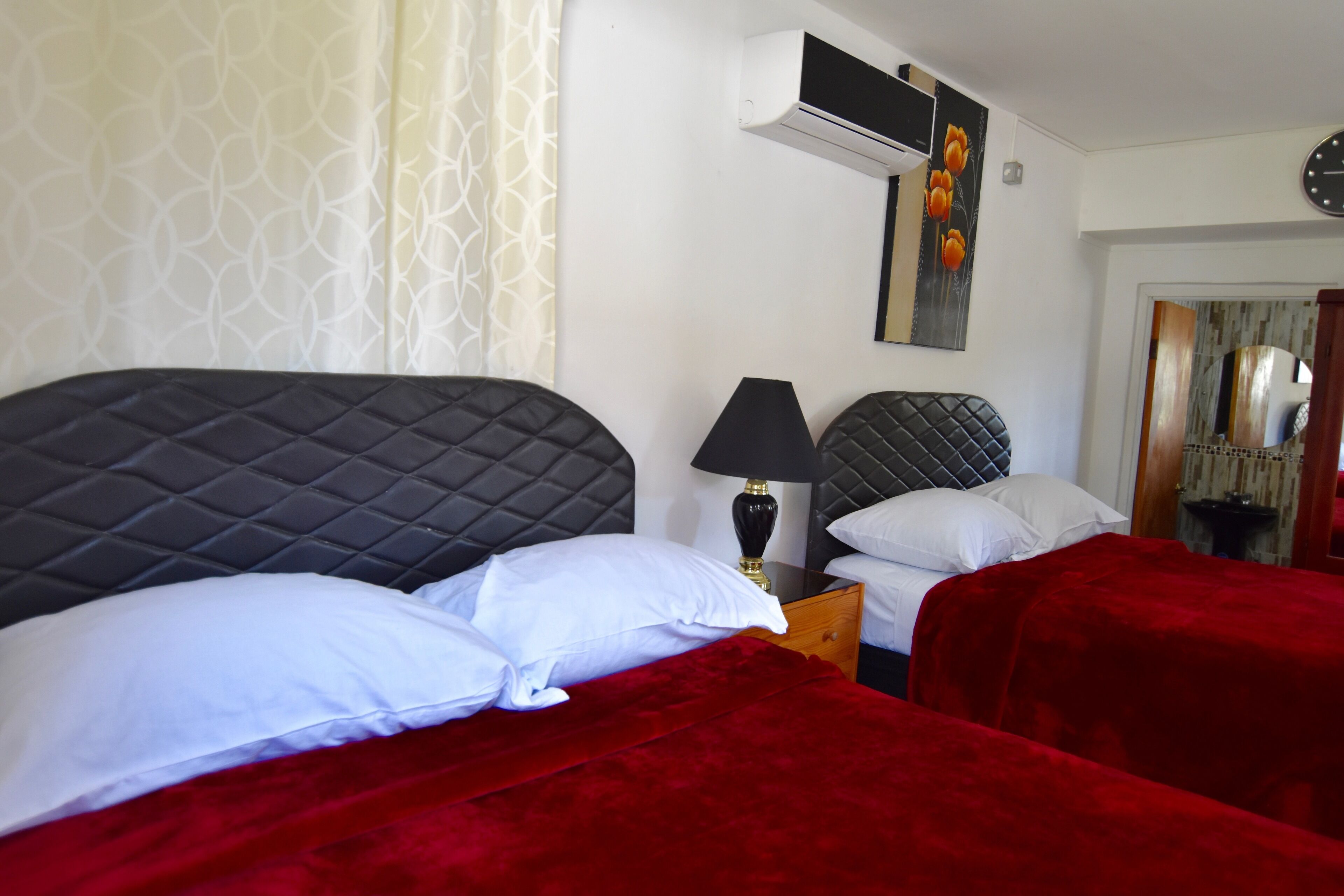 A hotel room with two beds, each with a black headboard and red bedding. There is a bedside table with a lamp, and an air conditioning unit mounted on the wall.