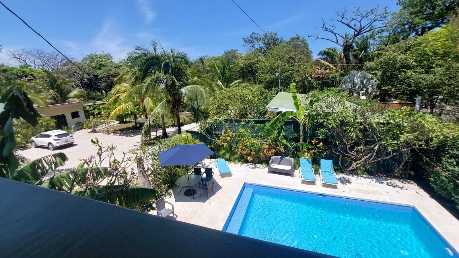 A pool area with a clear blue pool, lounge chairs, a table with chairs, and a car parked nearby.
