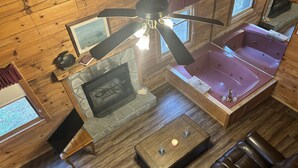 A cabin interior with a stone fireplace, a wooden ceiling fan, a stone-tub hot tub, a leather sofa, and a wooden coffee table. - SAVE!  ROMATIC  GATLINBURG   GETAWAY!!    (Gatlinburg)