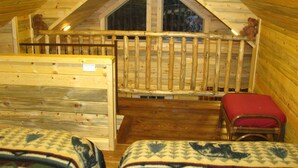 A cozy attic bedroom with a wooden sloped ceiling, a window, a wooden railing, a bench, and two beds with patterned blankets. - A little heavenly peace, cabin No. 2 (Polebridge)