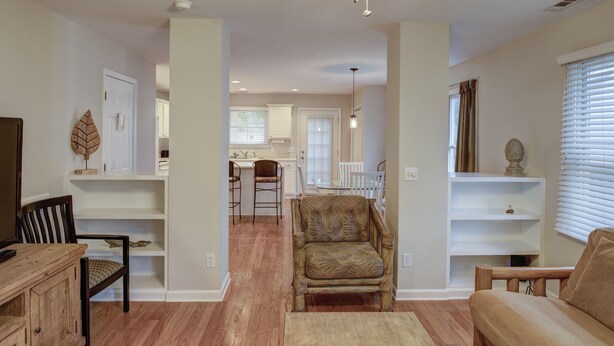 Un salón espacioso con suelo de madera, un sillón grande, un sofá y un mueble para televisión. Se observa una cocina al fondo con zona de comedor.