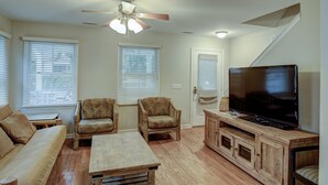 Una sala de estar con un sofá, dos sillones, una mesa de centro, un televisor sobre un soporte de madera y un ventilador de techo.