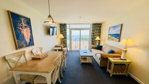 A hotel room with a dining table, chairs, a sofa, a small coffee table, and a view of the ocean through the balcony doors.