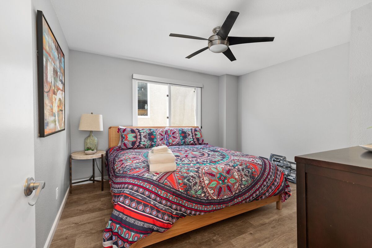 Een slaapkamer met een bed, een plafondventilator, een raam, een lamp en een dressoir.
