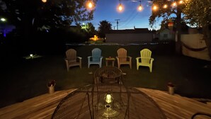Un área de estar al aire libre con luces colgantes, cuatro sillas Adirondack y una mesa metálica con un farol.