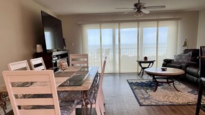 Ein Wohnzimmer mit Esstisch, Stühlen, einer Couch, einem Couchtisch, einem Fernseher und einem Balkon mit großen Fenstern.